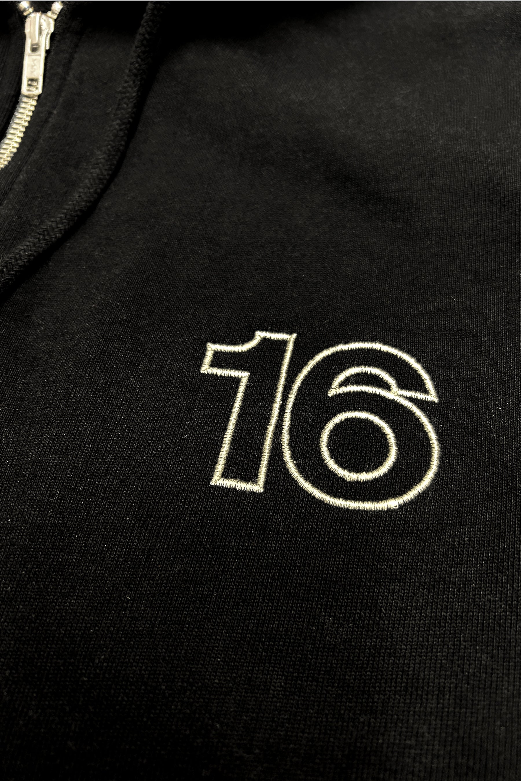 Close-up of 16 Sixteen embroidered logo on TRIBE WEAR zip-up hoodie